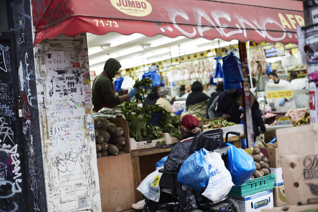 A busy fruit and veg shop entrance, woman in wheelchair, wearing a face mask with lots of shopping bags.