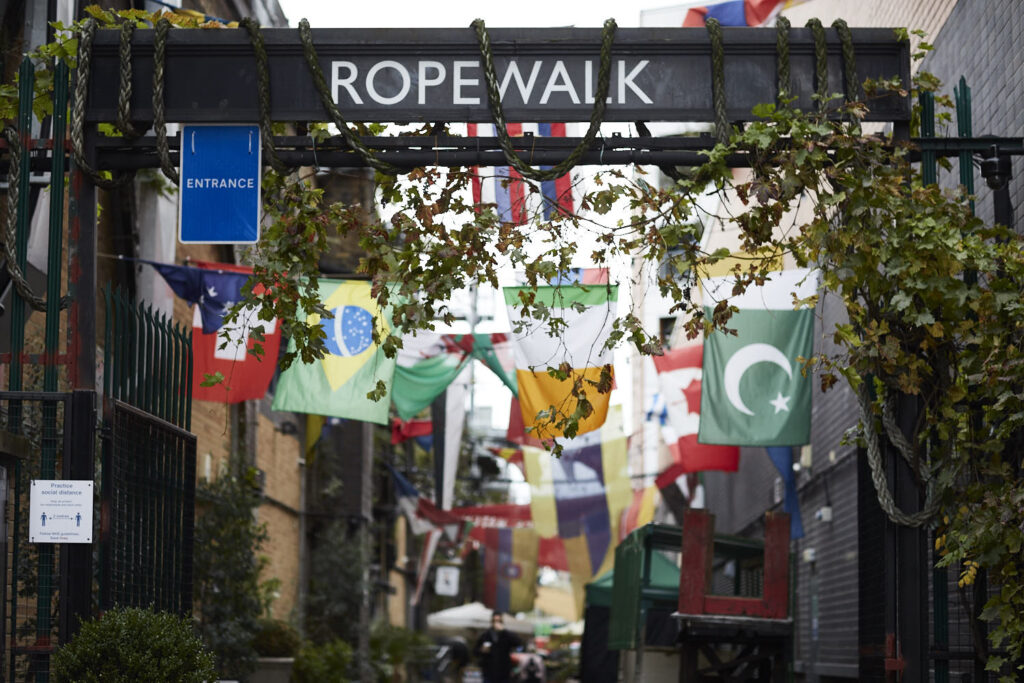 Rope Walk, South London. An ivy covered sign saying Rope Walk, with  lots of international flags hanging behind it.