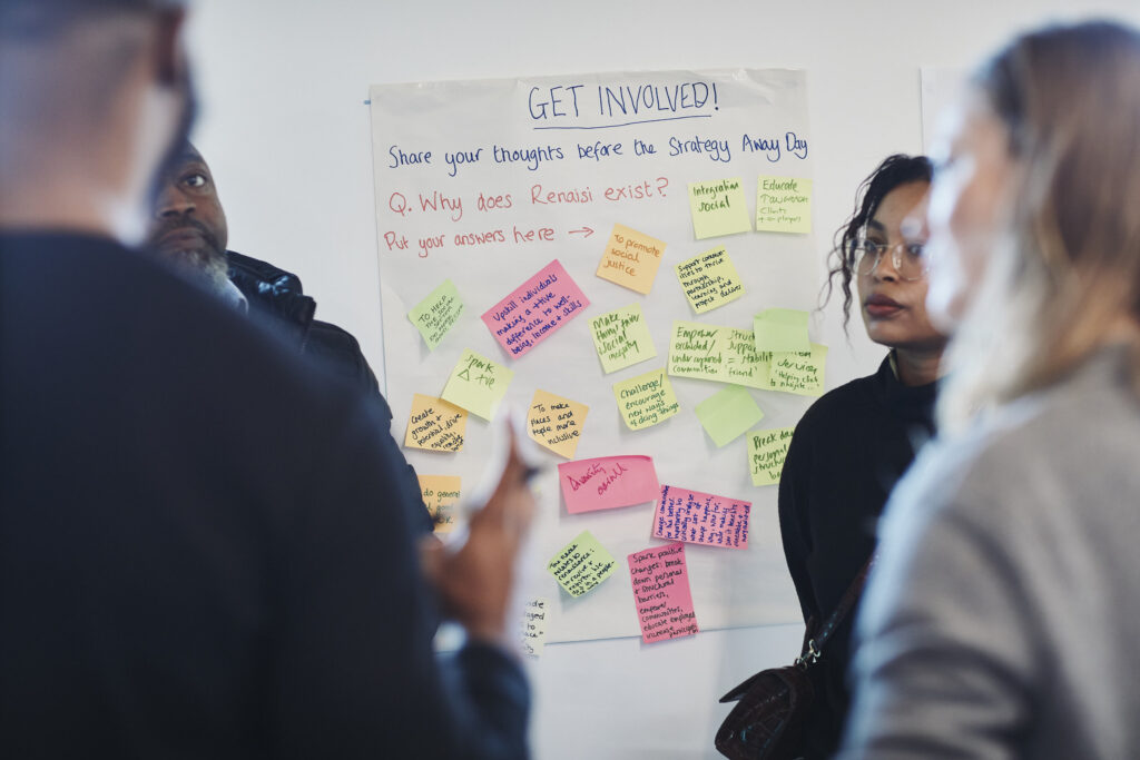 A group of Renaisi colleagues standing near a flipchart that reads "Why does Renaisi exist?" Answers on coloured post-it notes