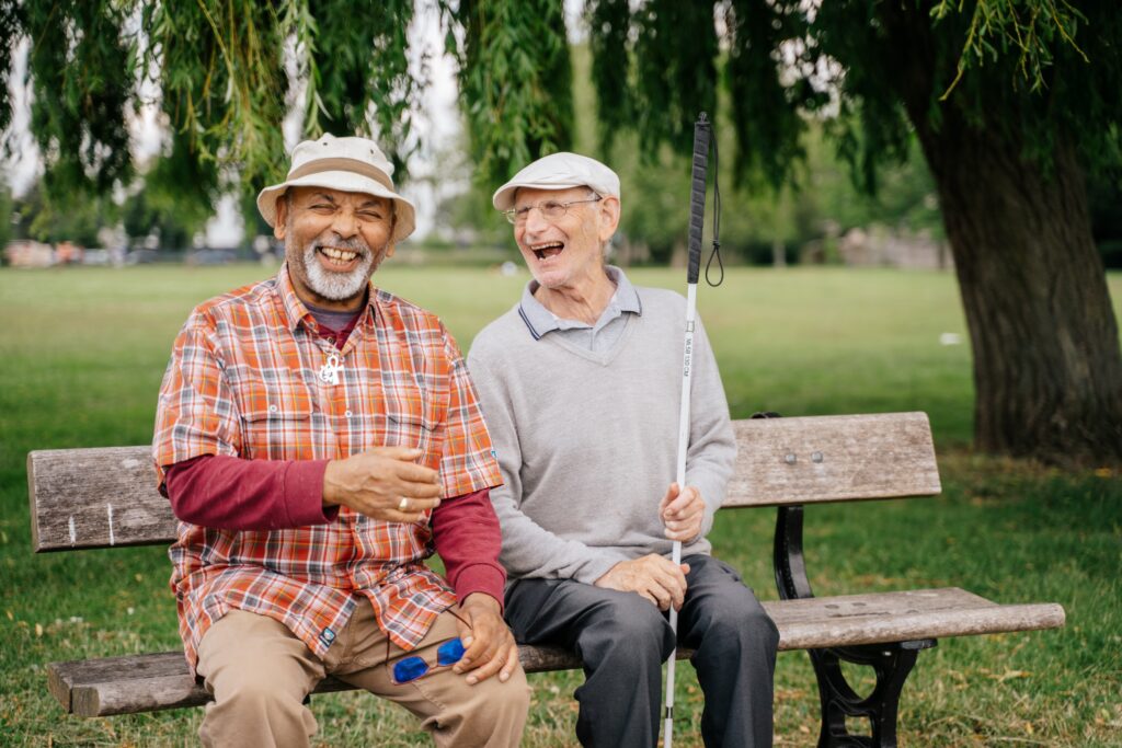 Two older men sitting on a bench, smiling and laughing together. One holds a mobility cane.